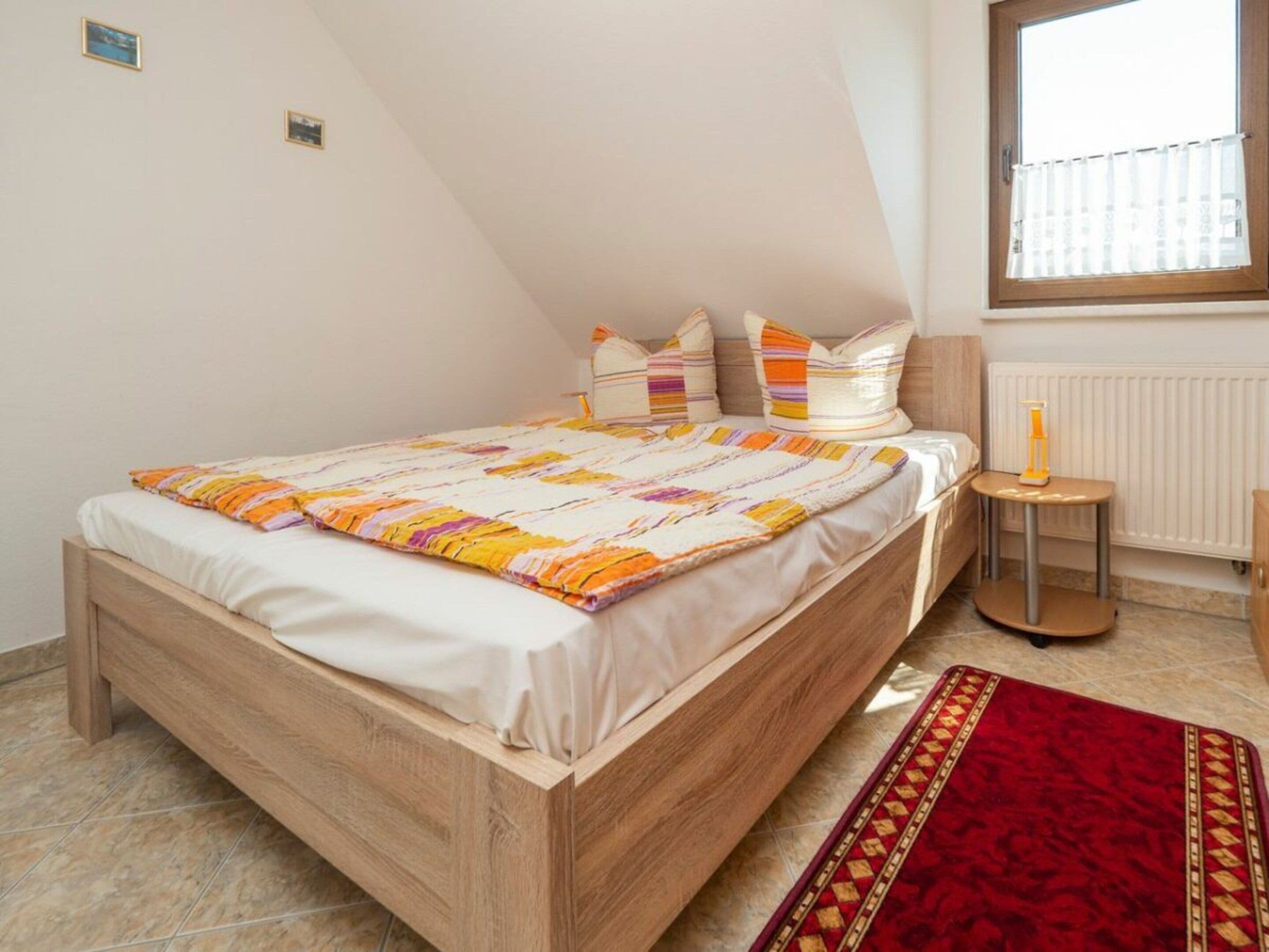 A bedroom with a wooden bed, a red rug, a small table with a bottle, and a window with blinds.