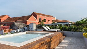Una piscina en la azotea con cubierta de madera y sillones de descanso, rodeada de edificios con tejados de tejas rojas.