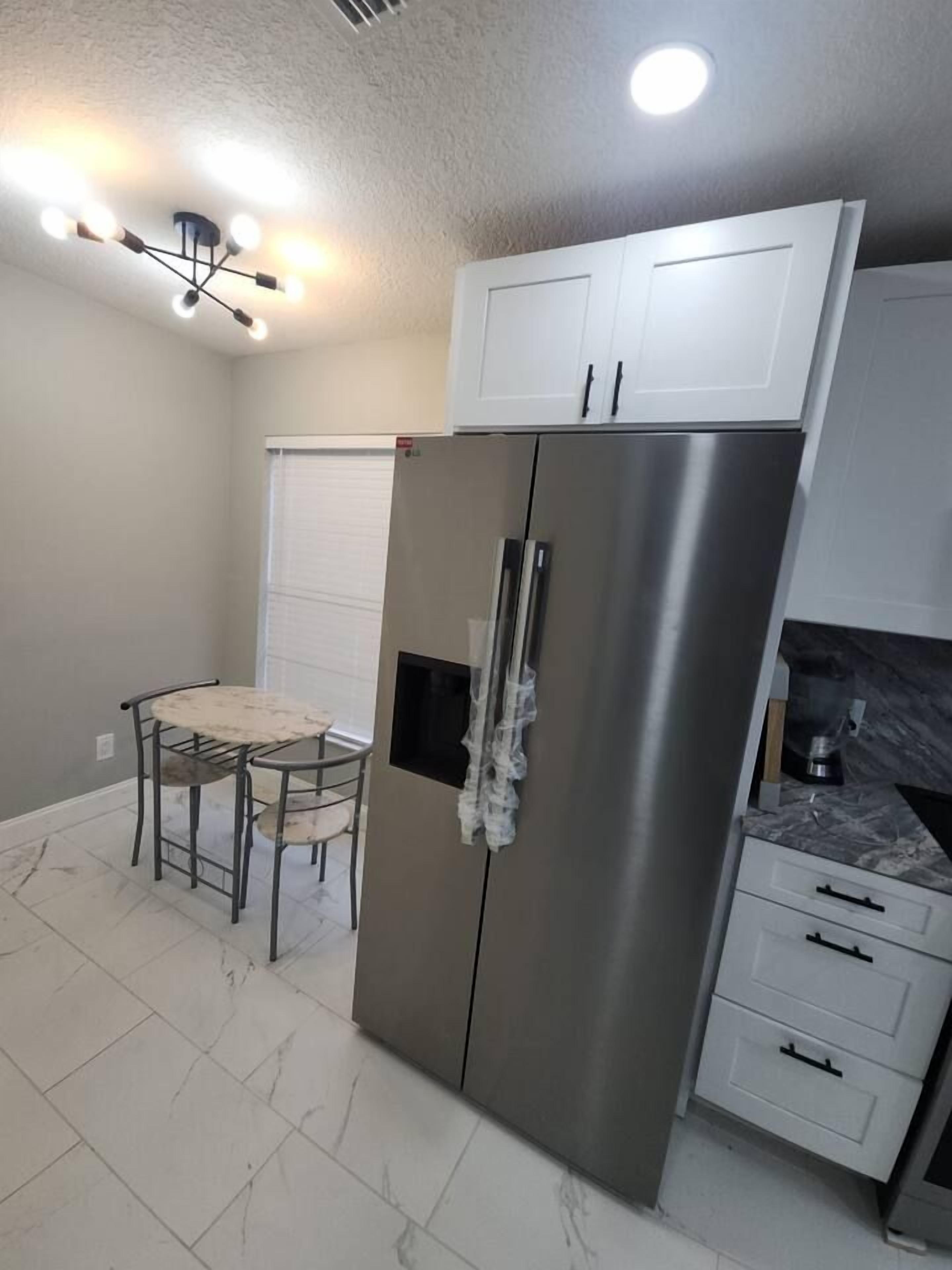 A compact kitchen with a stainless steel refrigerator, white cabinets, a small dining table with chairs, and a ceiling light fixture.