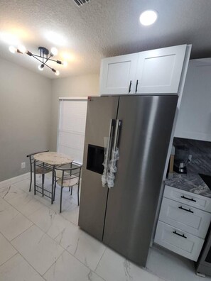 A compact kitchen with a stainless steel refrigerator, white cabinets, a small dining table with chairs, and a ceiling light fixture. - Modern Tampa Retreat Hot Tub  EV Charger Sleeps 8 (Tampa)