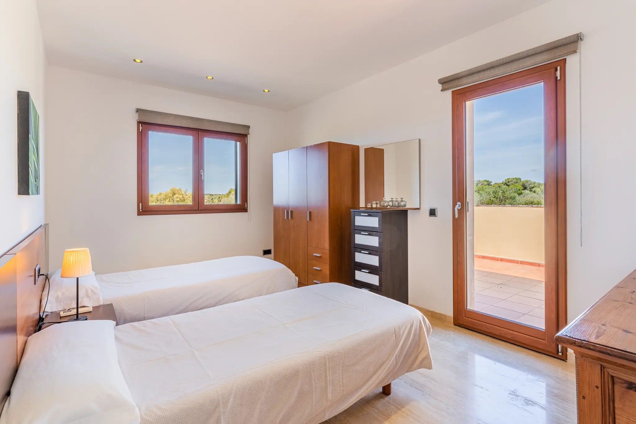 A bedroom with two beds, a wardrobe, a desk, a lamp, and a window with a view.
