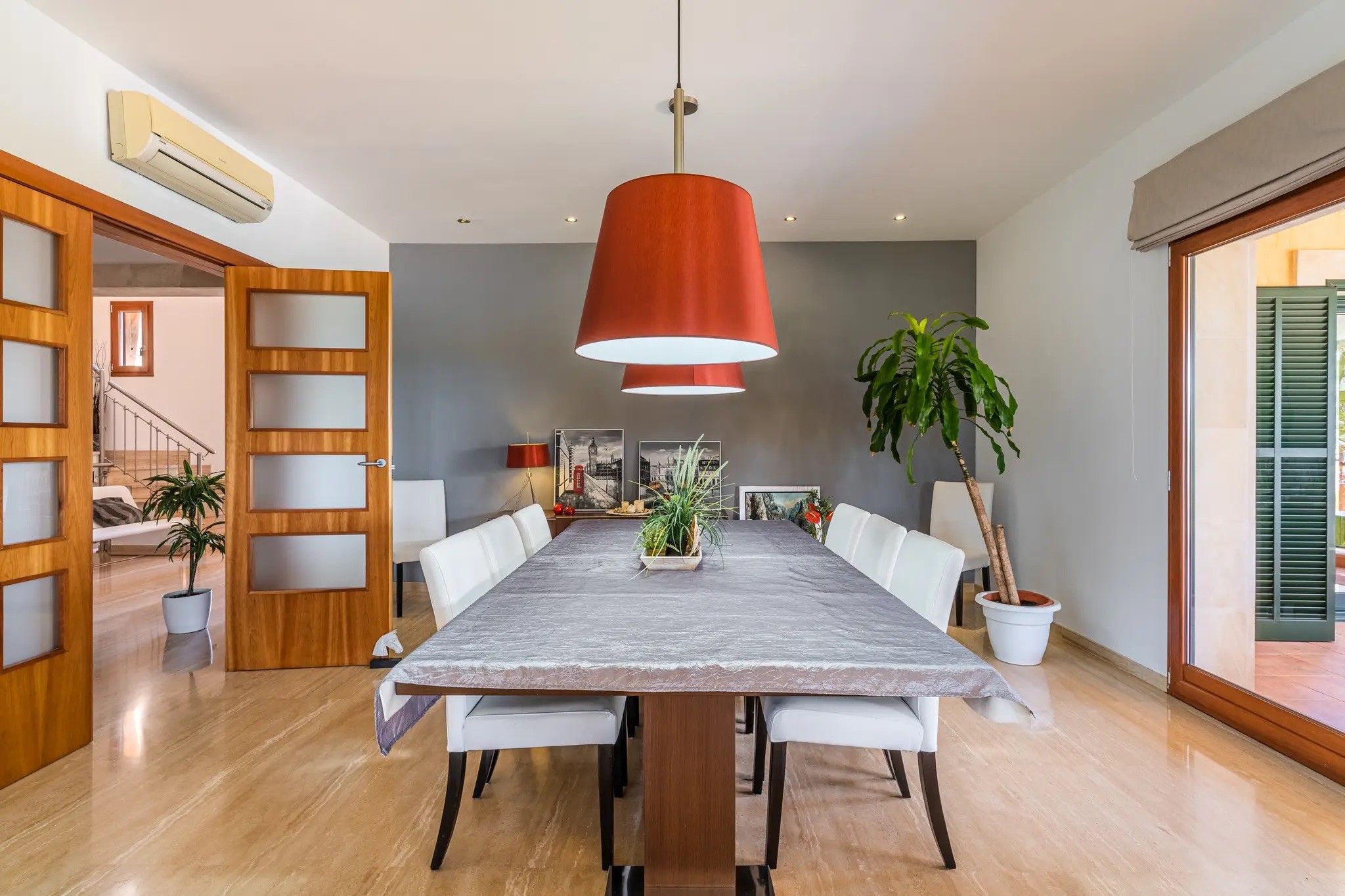 A dining area with a long table, white chairs, a red pendant light, and potted plants.