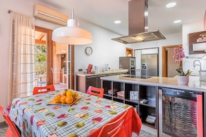 A modern kitchen with a dining area, featuring a fruit bowl on the table, a coffee maker, and a refrigerator. - Holiday Home 'Can Bou' with Sea View, Wi-Fi and Air Conditioning (Ses Salines)