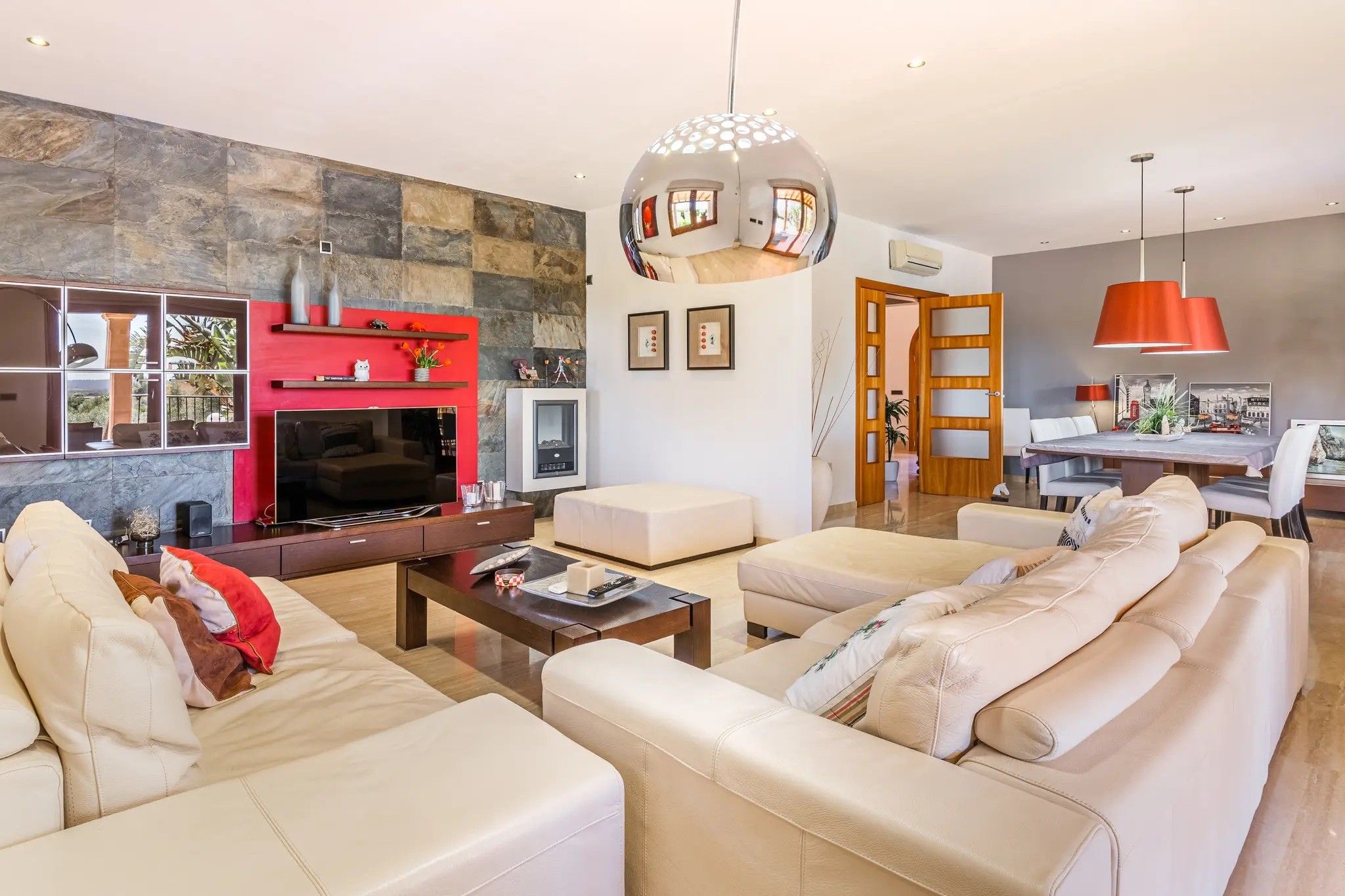 Una sala moderna con chimenea, un sofá grande, una mesa de centro y un comedor con una lámpara colgante roja.
