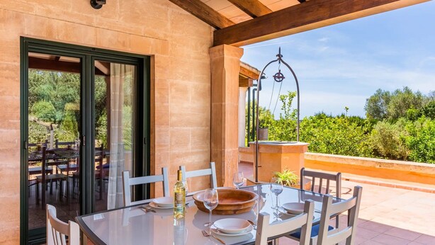 Mesa com tampo de vidro, cadeiras, garrafa e copos de vinho em um pátio com vista para vegetação e uma pérgola de madeira.
