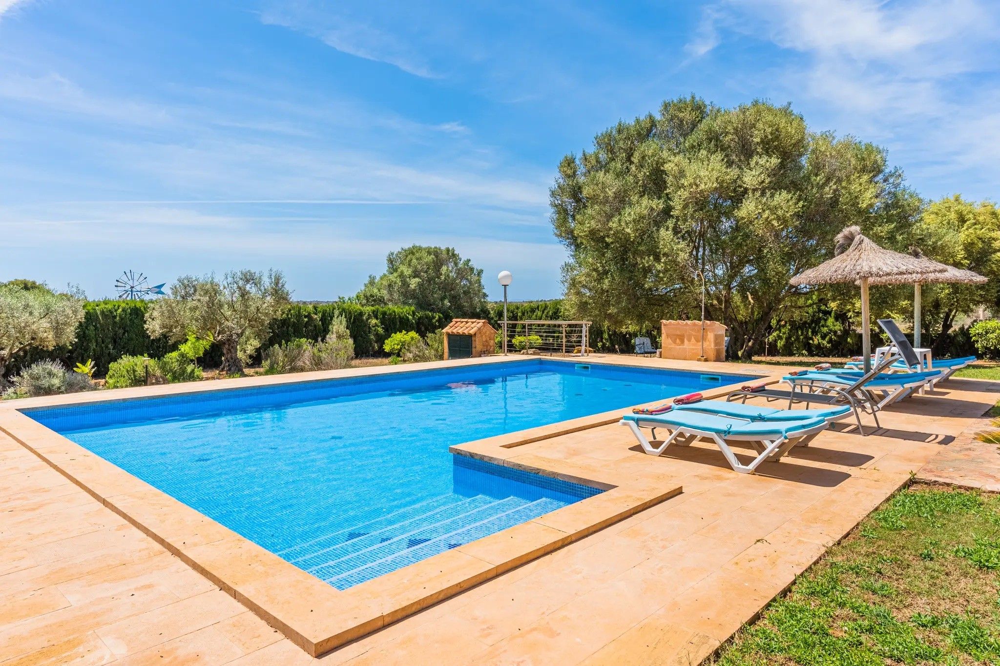 Una piscina rectangular con borde de azulejos, rodeada por un solárium empedrado. Se observan sillones de descanso y un pequeño pabellón.