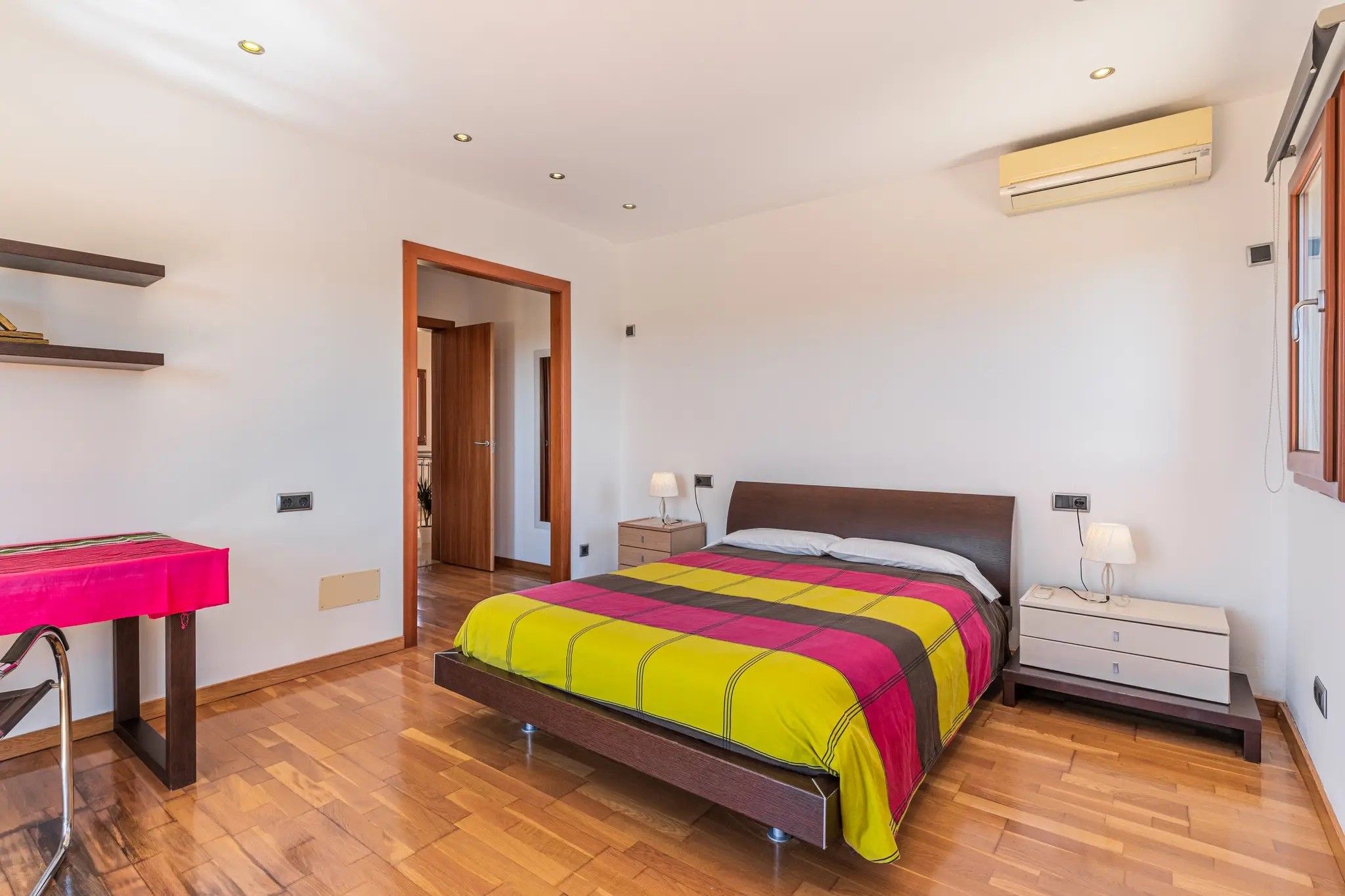 A bedroom with a bed, bedside tables, an air conditioning unit, a desk, and a chair.
