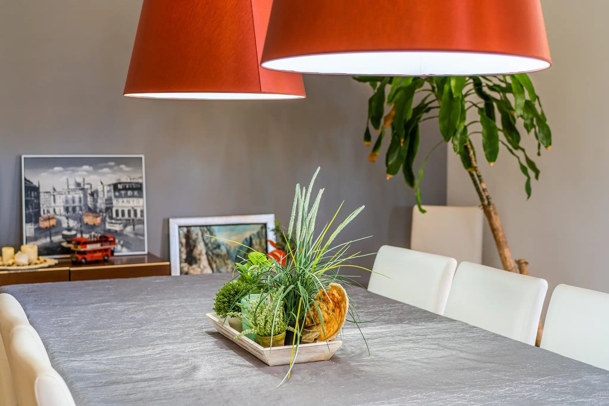Een eettafel met een bloemstuk in het midden, een lijst met foto aan de muur en een lamp.