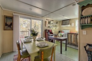 A dining area with a table set for four, a kitchen in the background, and a window with a view of trees. - Cozy Mountain Cottage w/ Custom Laurel Furnishings — Pet Friendly, Sleeps 4 (Lakemont)