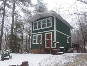 A two-story cabin with a red door, surrounded by a snowy forest. - Cozy Mountain Cottage w/ Custom Laurel Furnishings — Pet Friendly, Sleeps 4 (Lakemont)