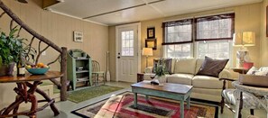 A cozy living room with a sofa, coffee table, and a rustic wooden chair. A bookshelf and a small table with a bowl of fruit are also visible. - Cozy Mountain Cottage w/ Custom Laurel Furnishings — Pet Friendly, Sleeps 4 (Lakemont)