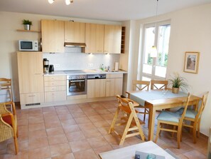 A compact kitchen with wooden cabinets, a microwave, a stove, and a sink. There is a dining area with a table and chairs. - Apartment Building Linquenda 09 (Heringsdorf)