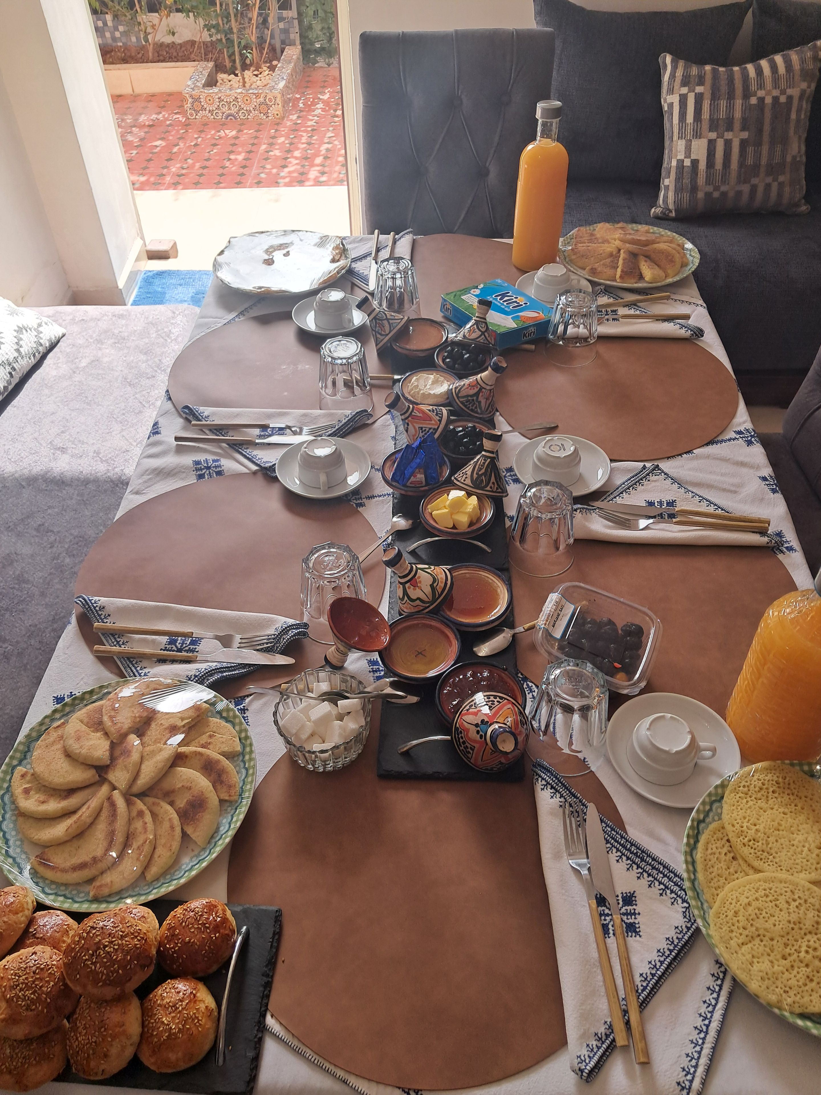 Une table à manger dressée avec différents plats, couverts et une bouteille de jus d’orange.