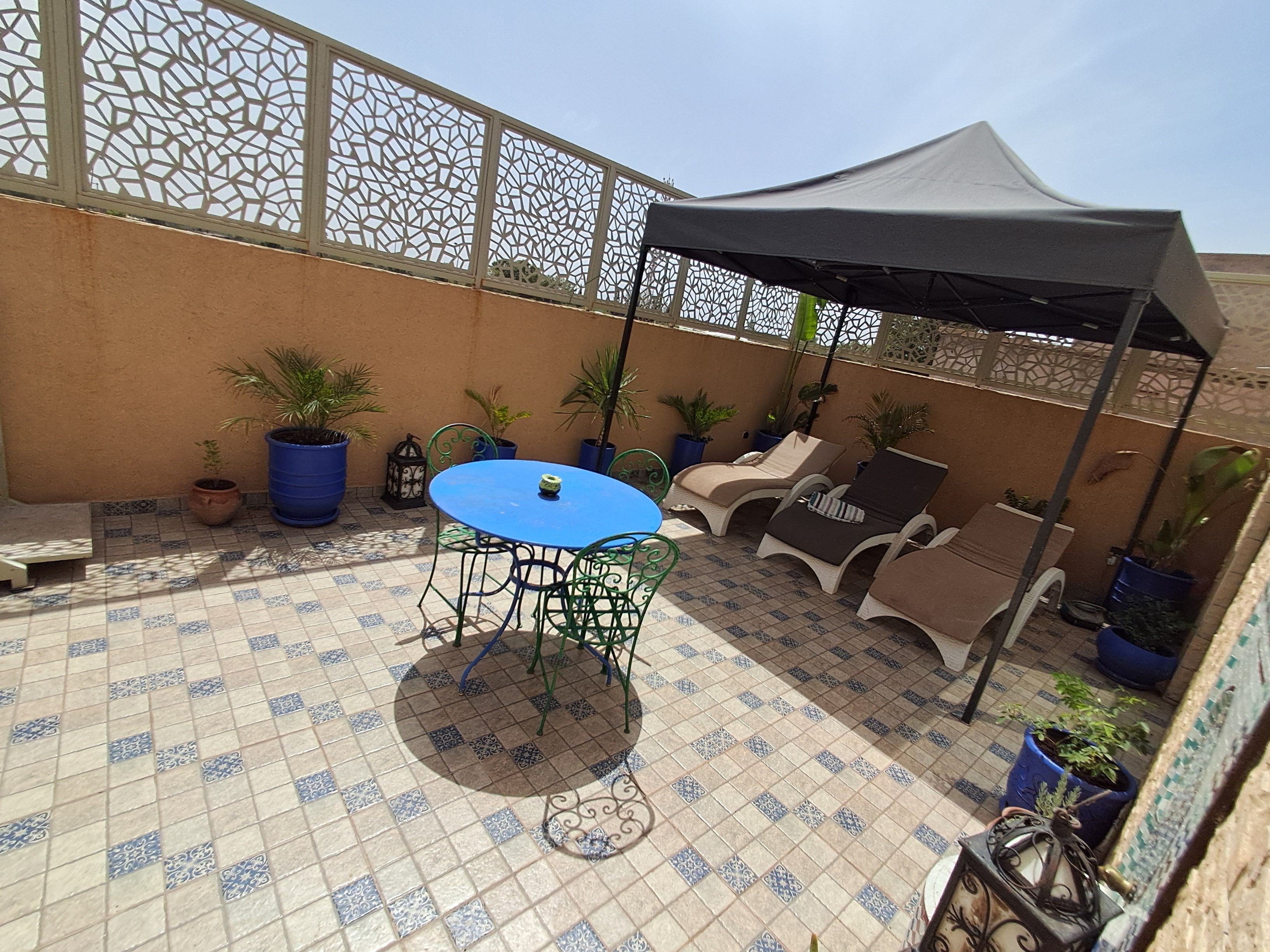 An outdoor patio with a blue table and chairs, potted plants, and a covered area with lounge chairs.