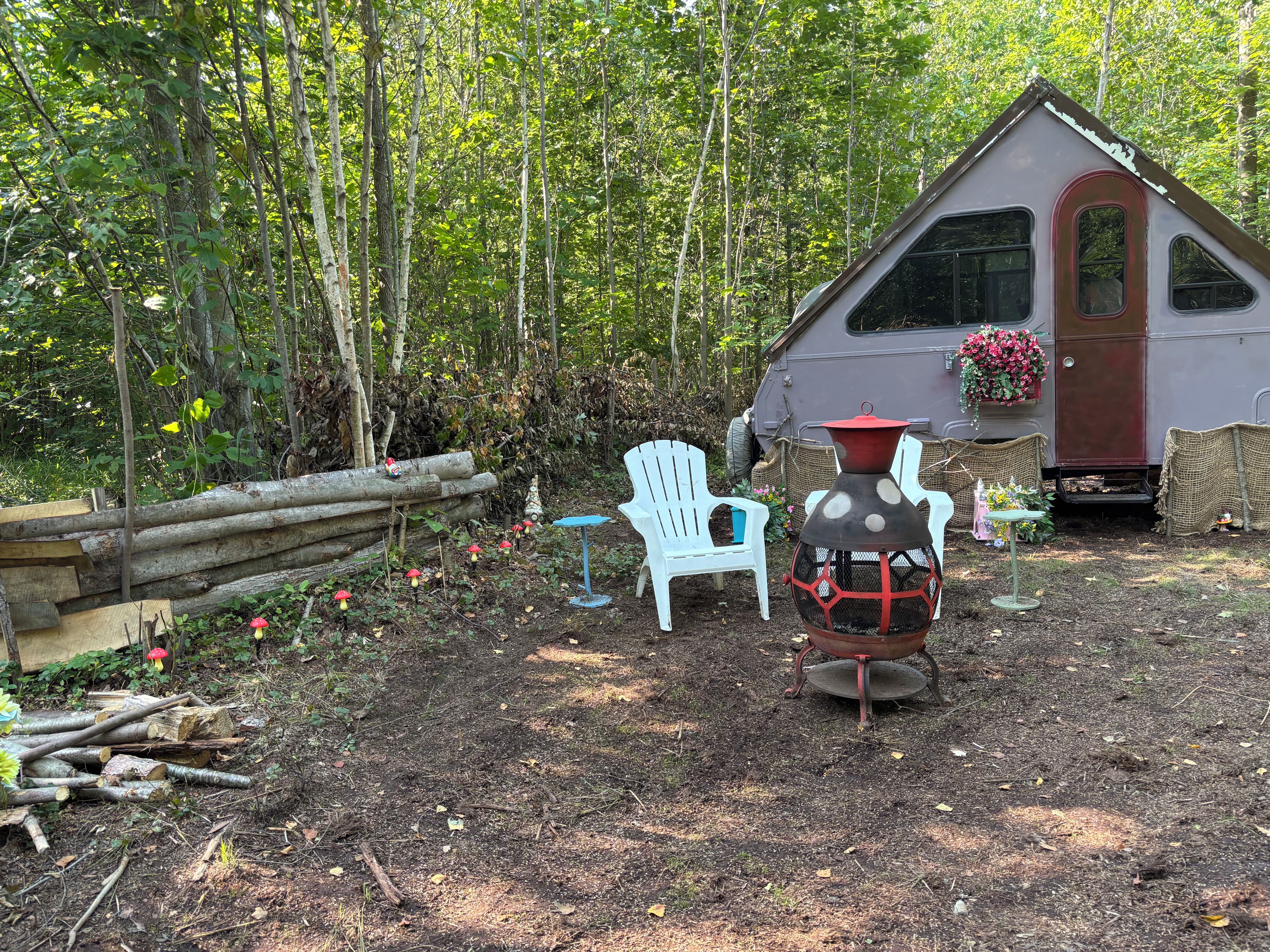 Un chalet rustique avec une cheminée, entouré d’une forêt. On y trouve deux chaises blanches et une petite table agrémentée de fleurs.