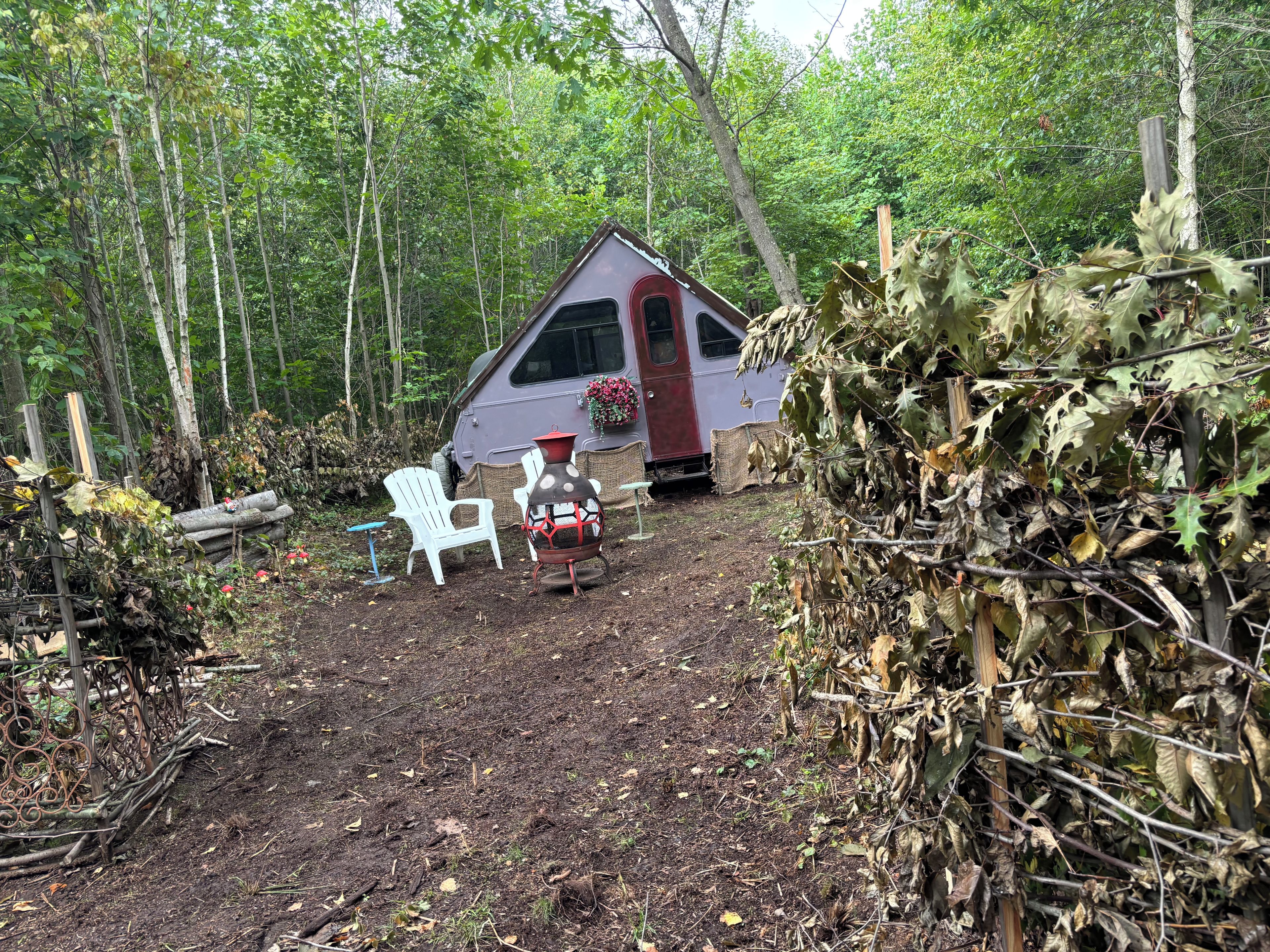 Une cabane rustique avec une porte rouge, entourée d’une forêt. On y trouve des chaises blanches et une lanterne à l’extérieur.