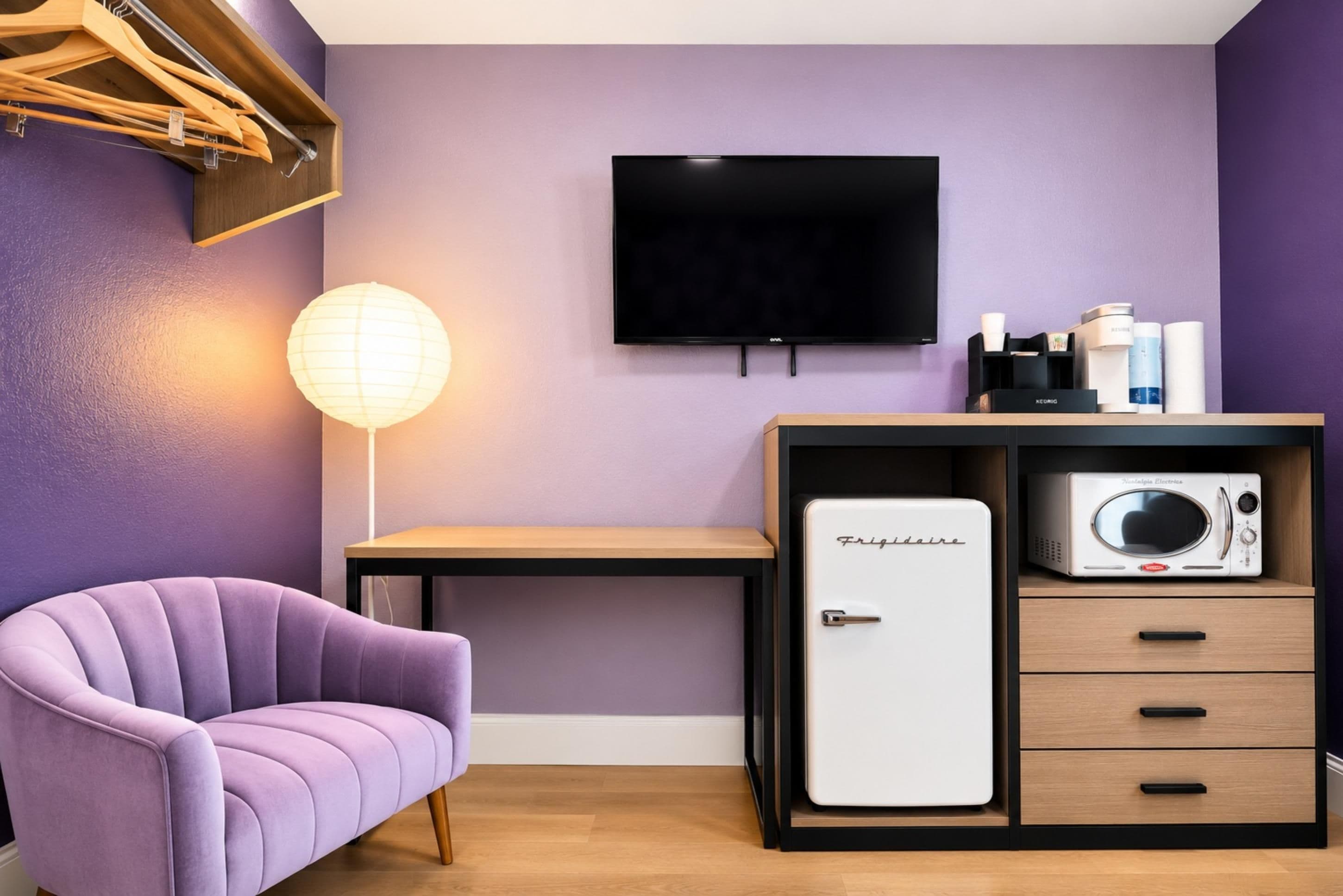 Quarto com parede roxa, TV de tela plana, geladeira estilo vintage, escrivaninha de madeira, cadeira roxa e luminária de chão.