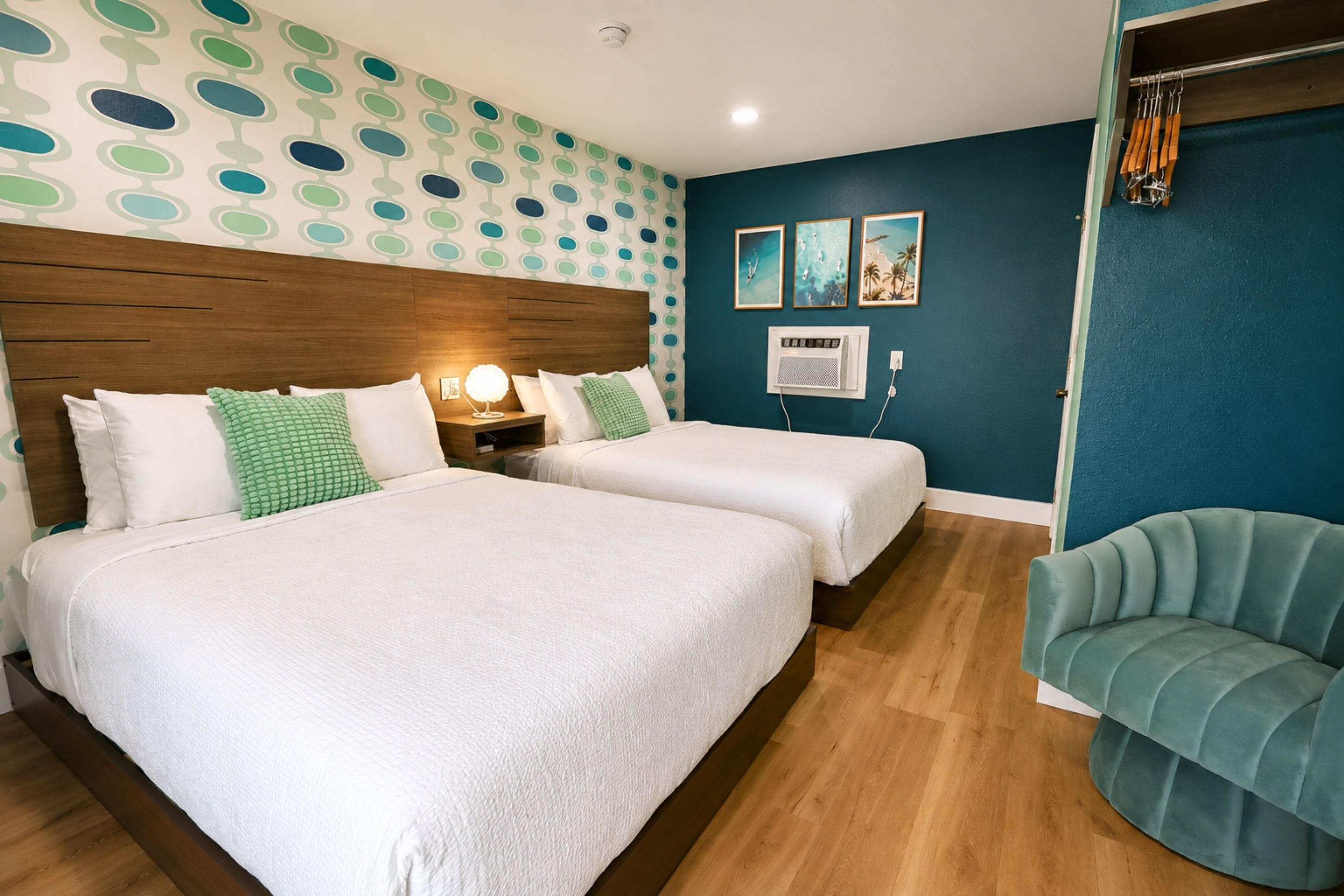 Habitación de hotel con dos camas, cabecero de madera, pared de acento color aguamarina y un pequeño rincón de descanso.
