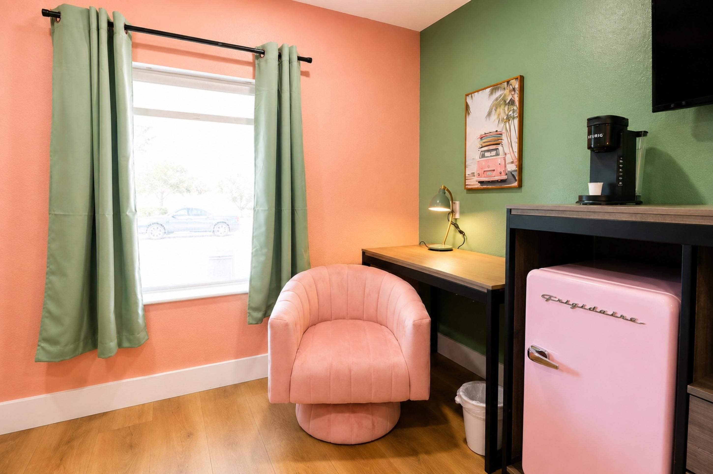 Una habitación con una silla rosa, un refrigerador, un escritorio con una lámpara y una ventana con cortinas.