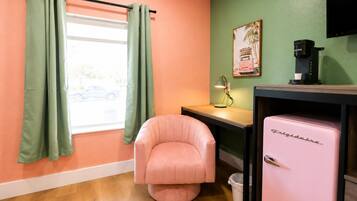 Una habitación con una silla rosa, un refrigerador, un escritorio con una lámpara y una ventana con cortinas.