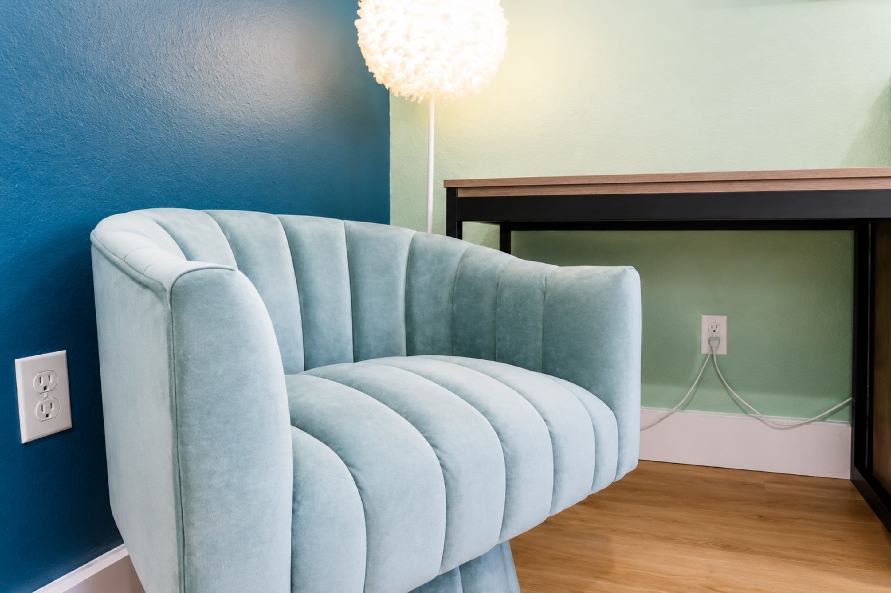 A room with a blue wall, a light blue upholstered chair, a wooden table, and a white electrical outlet.