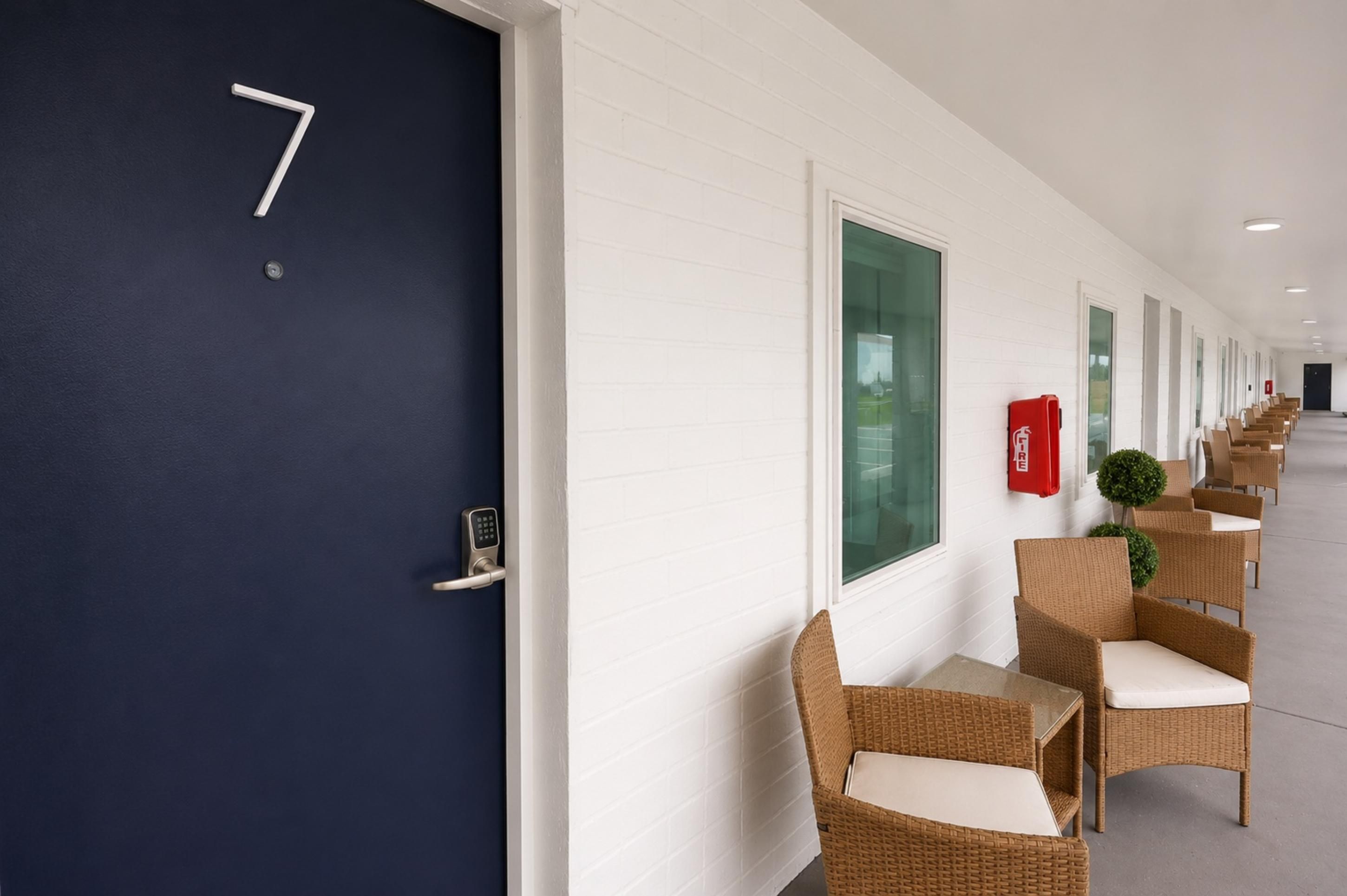 Een gang met een donkerblauwe deur met het nummer 7, witte muren en rieten stoelen met witte kussens.