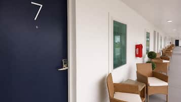 Een gang met een donkerblauwe deur met het nummer 7, witte muren en rieten stoelen met witte kussens.