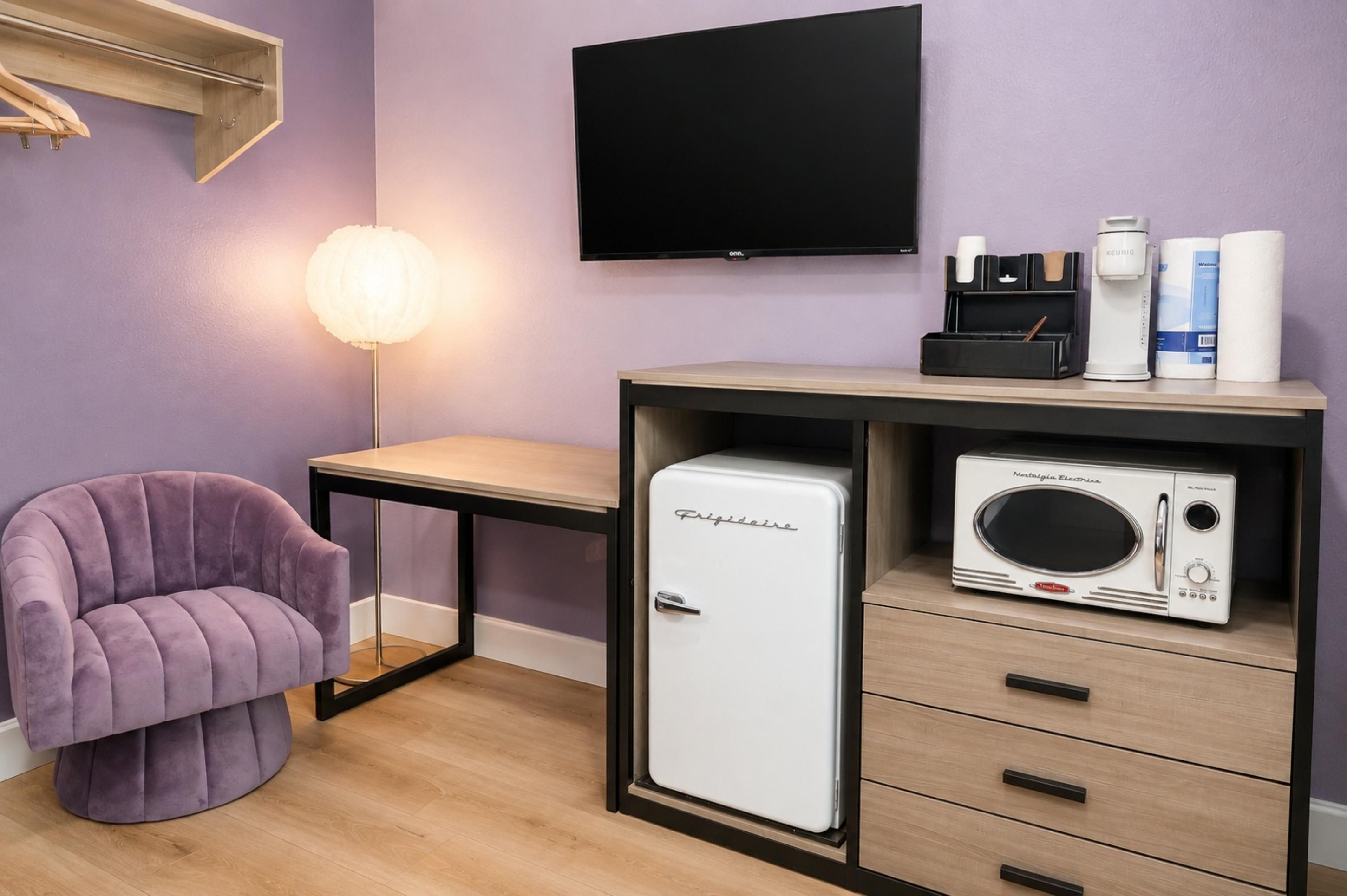 Een compacte kamer met een paarse wand, een flatscreen televisie, een kleine witte koelkast, een magnetron, een houten tafel, een paarse stoel en een bedlamp.