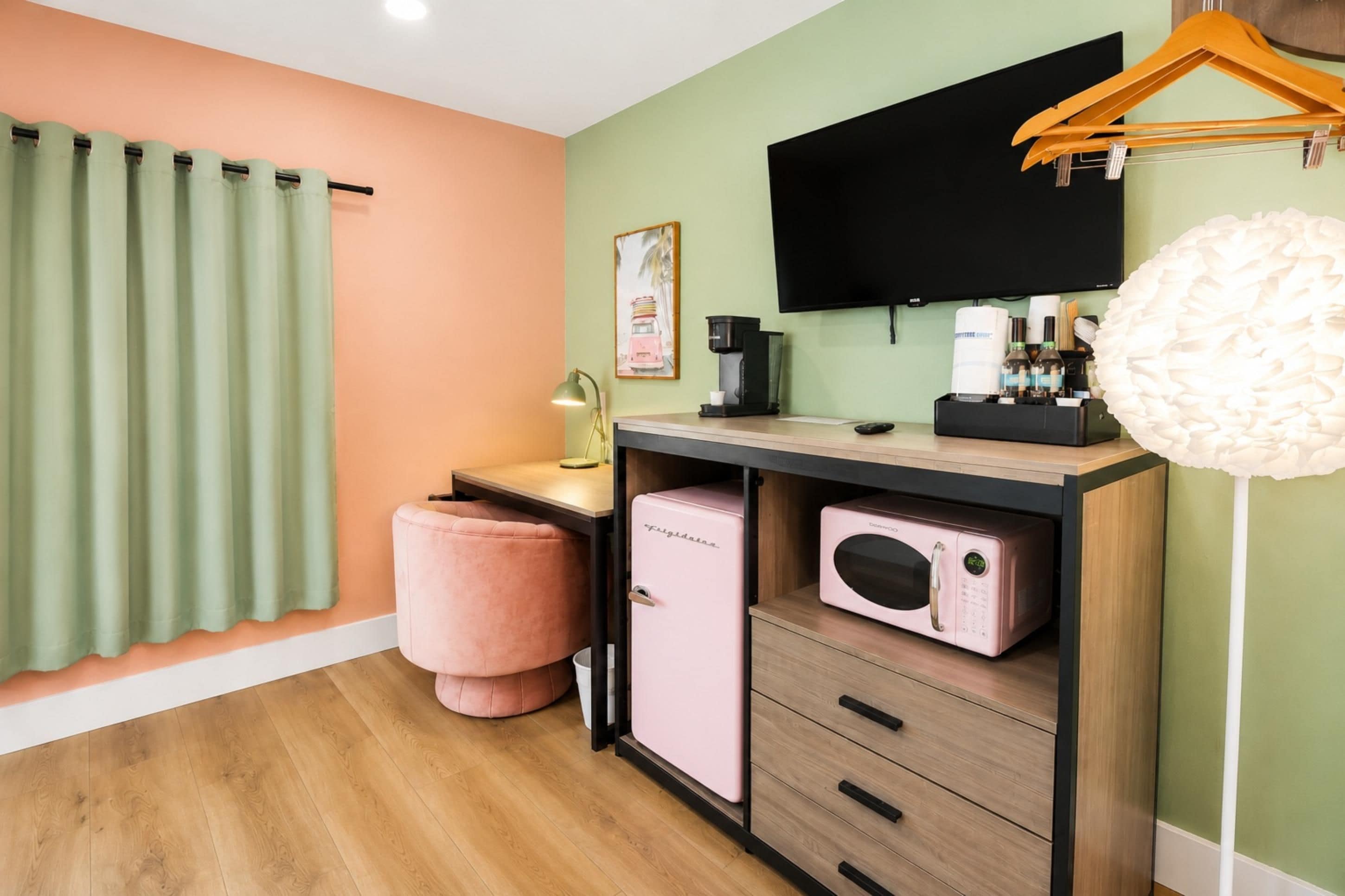 A room with a wooden desk, a pink chair, a television mounted on the wall, and a coffee machine.