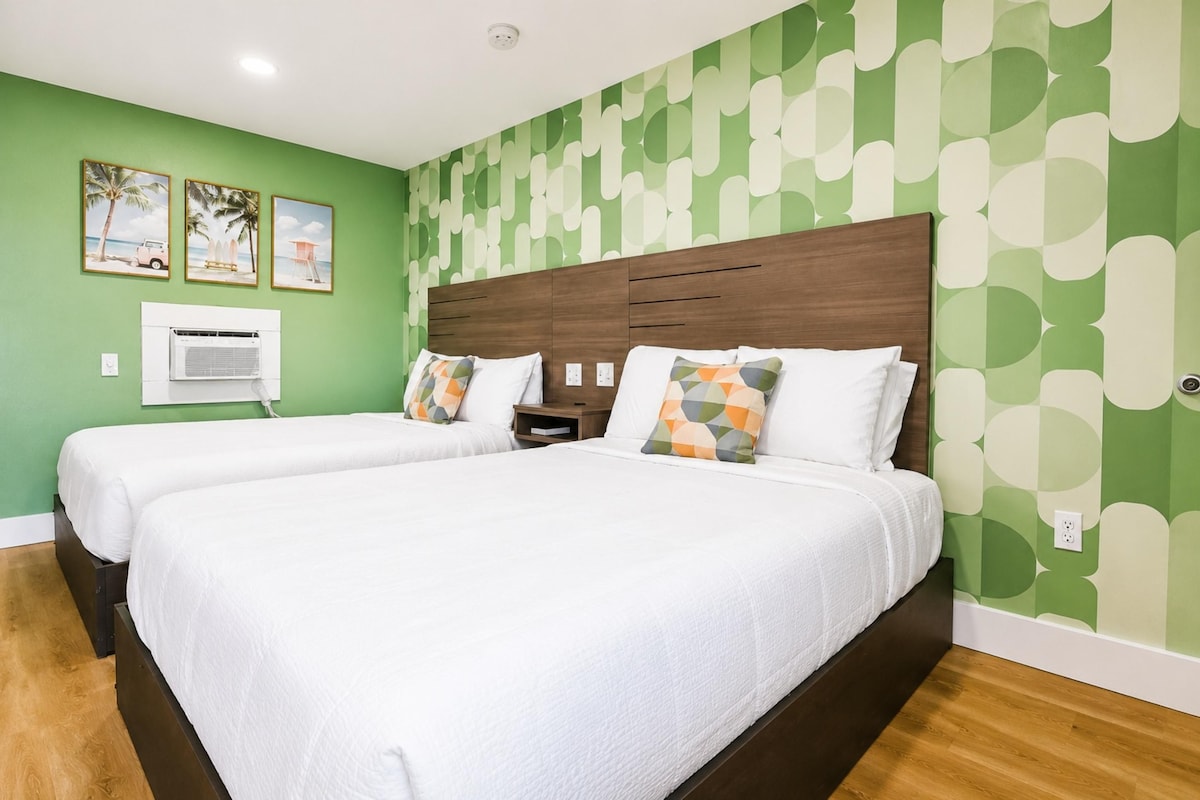 A hotel room with two beds, a wooden headboard, a wall-mounted air conditioner, and a beach-themed picture on the wall.