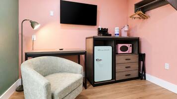 Una habitación con escritorio, una silla, kitchenette con refrigerador, microondas y televisor montado en la pared.