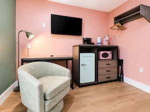 Una habitación con escritorio, una silla, kitchenette con refrigerador, microondas y televisor montado en la pared.