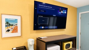 A room with a mounted TV displaying a smart hub interface, a framed picture of a beach scene, a coffee machine, a mini fridge, and a coffee bar. - The Blu - A Boutique Motel (Port St. Joe)