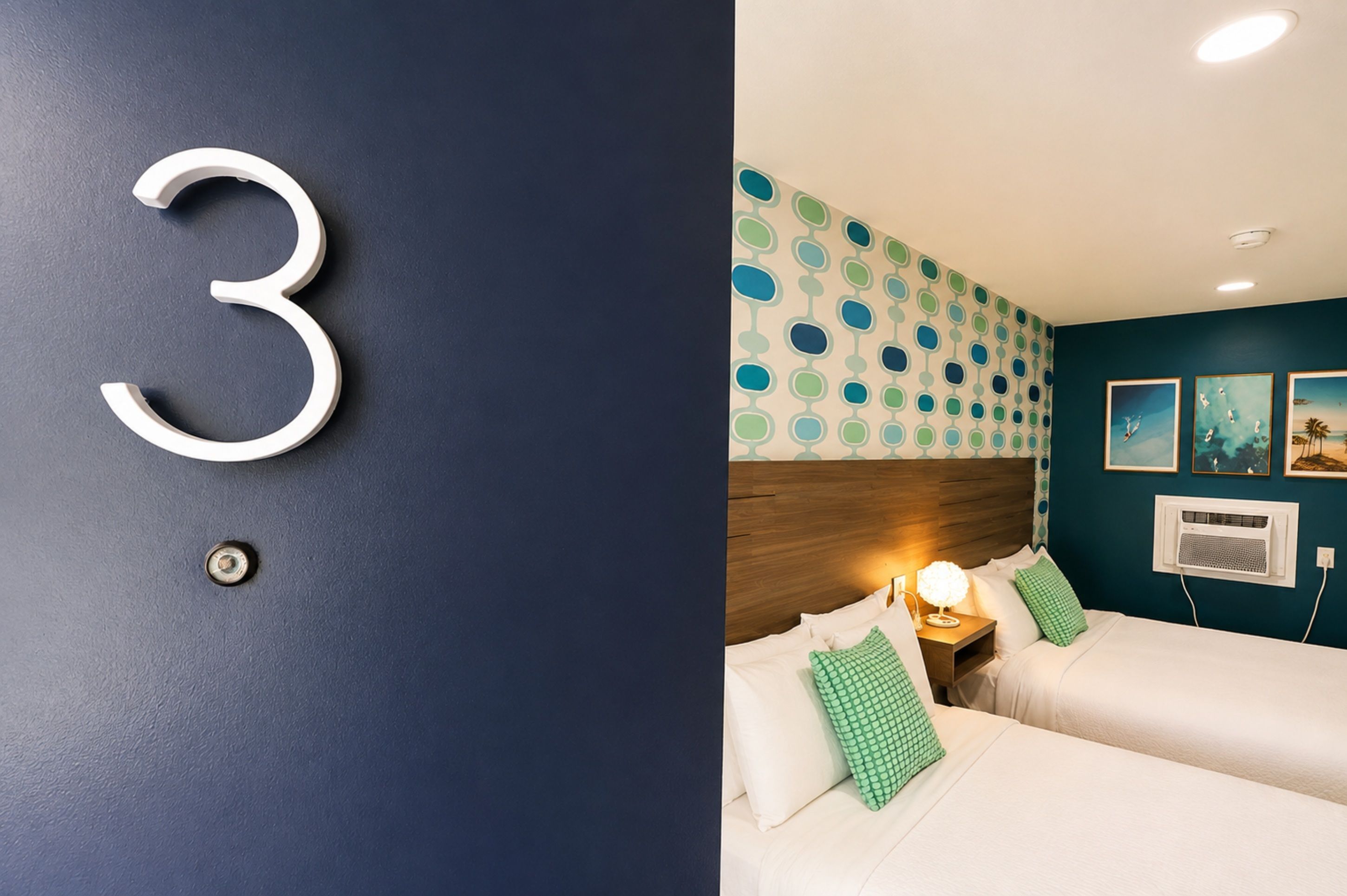 A hotel room with a large number '3' on the wall, a patterned wallpaper, a wooden headboard, two beds with white bedding, green pillows, a nightstand with a lamp, and a wall-mounted air conditioning unit.