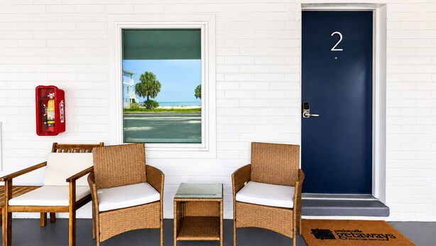 A room with two wicker chairs, a small table, a fire extinguisher, a door with the number 2, and a window with a view of a building and palm trees. - The Blu - A Boutique Motel (Port St. Joe)