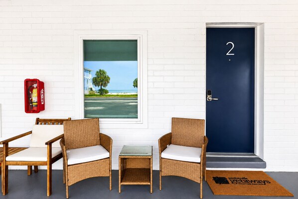 A room with two wicker chairs, a small table, a fire extinguisher, a door with the number 2, and a window with a view of a building and palm trees. - The Blu - A Boutique Motel (Port St. Joe)