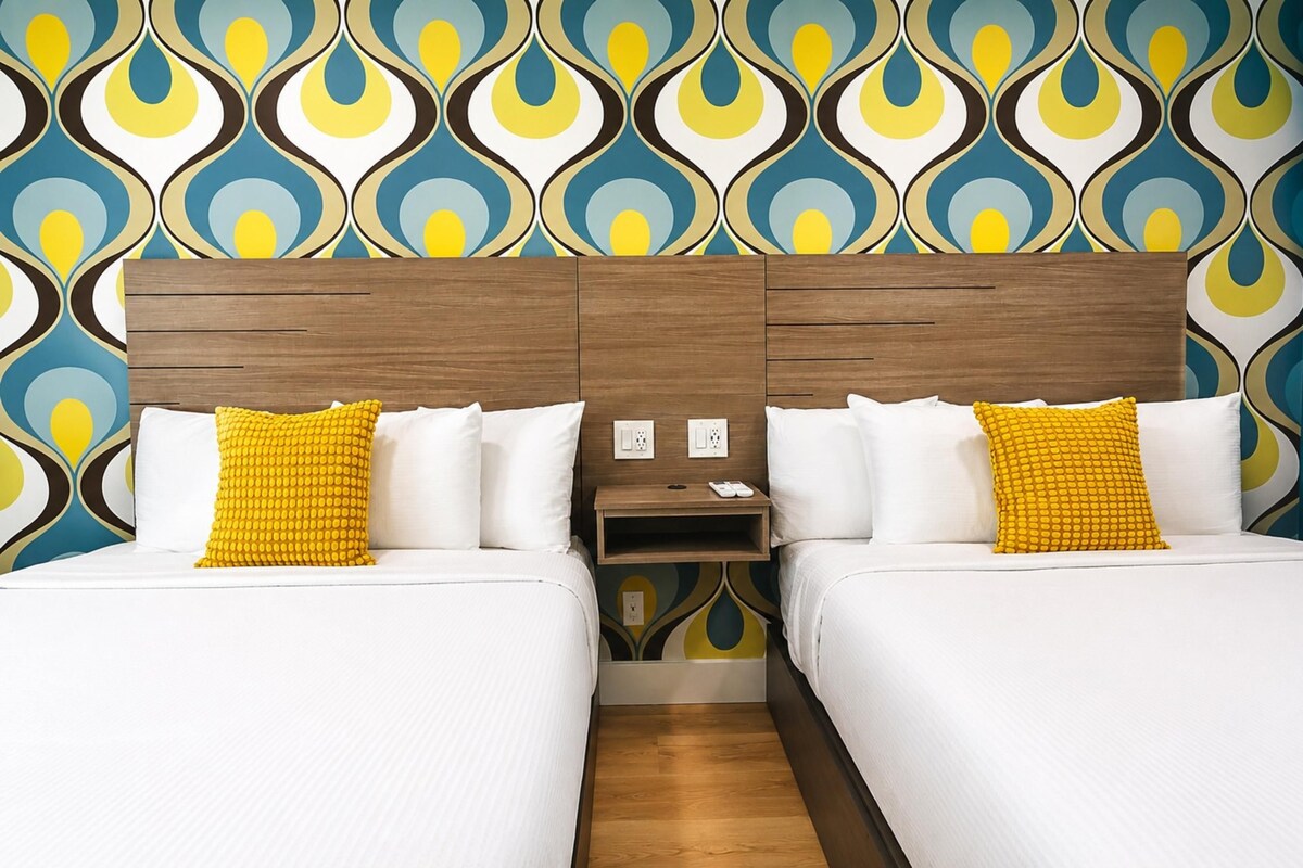 A hotel room with two beds, a wooden headboard, a nightstand, and a patterned wallpaper.