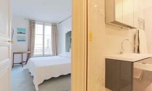 A hotel room with a bed, a window with curtains, a small table, and a bathroom with a sink and mirror. - LUXURIOUS 145M² APARTMENT ON RUE FRANÇOIS 1ER NEAR CHAMPS-ÉLYSÉES (Paris)