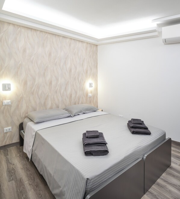A modern bedroom with a bed, two bedside lamps, a wall-mounted air conditioner, and a wooden floor.