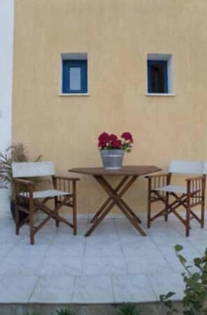 An outdoor seating area with a table and chairs, a potted plant, and a building with two windows. - Mourayio '' 'Jasmine' ' (Paphos)