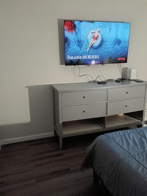 A television mounted on a wall displaying 'The Paradise Murders' on Netflix, a grey dresser with drawers, and a bed with a blue blanket. - 2-bedroom apartment in charming Atlanta with cool AC and gym access (Atlanta)