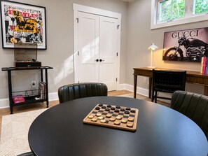 Ein Zimmer mit einem runden Tisch, einem Schachbrett, einem gerahmten Beatles-Poster, einem gerahmten Gucci-Motorrad-Poster und einem Schreibtisch mit einer Lampe.