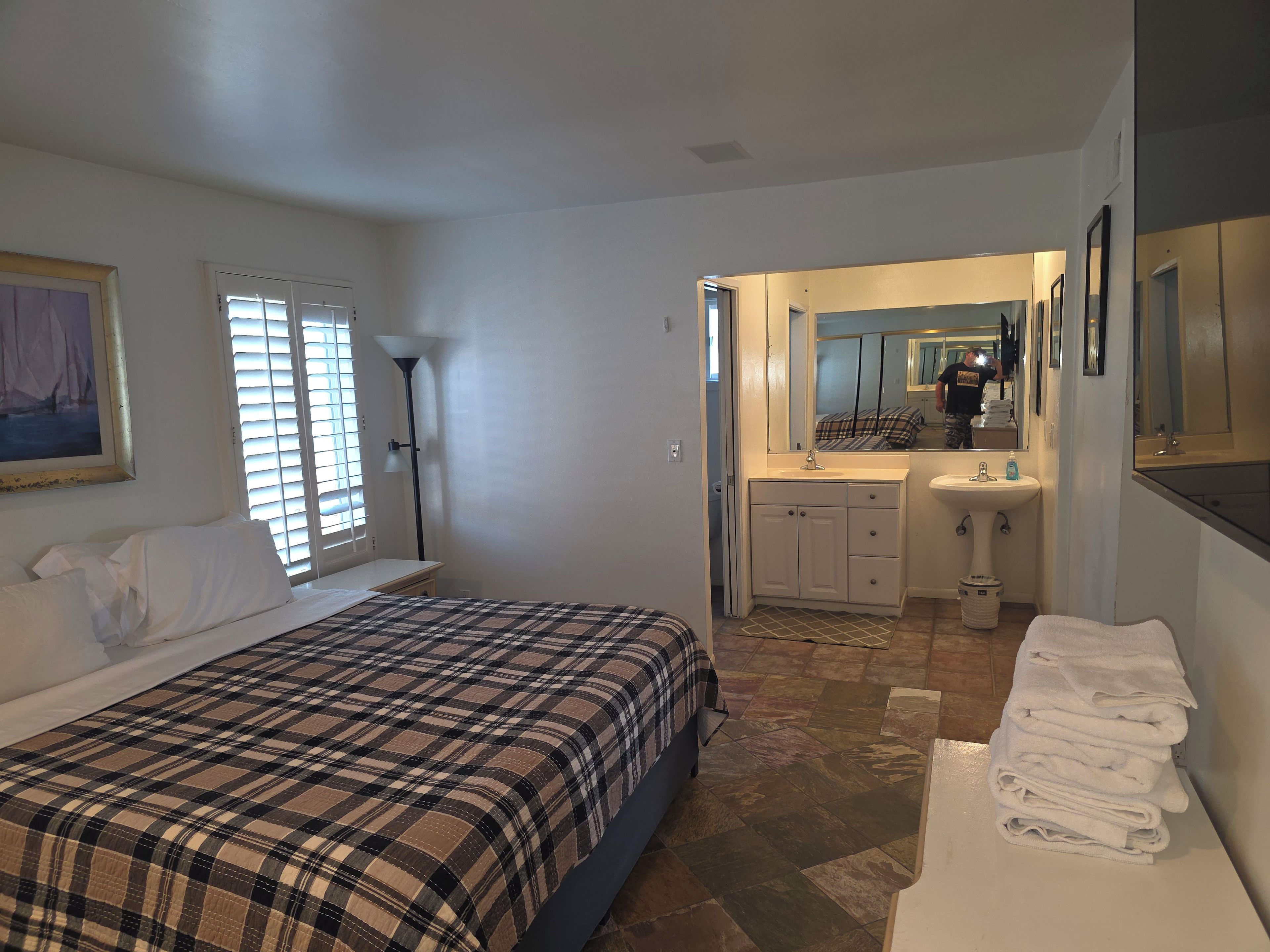 Quarto com cama, mesa de cabeceira, luminária, janela com persianas e pintura na parede.