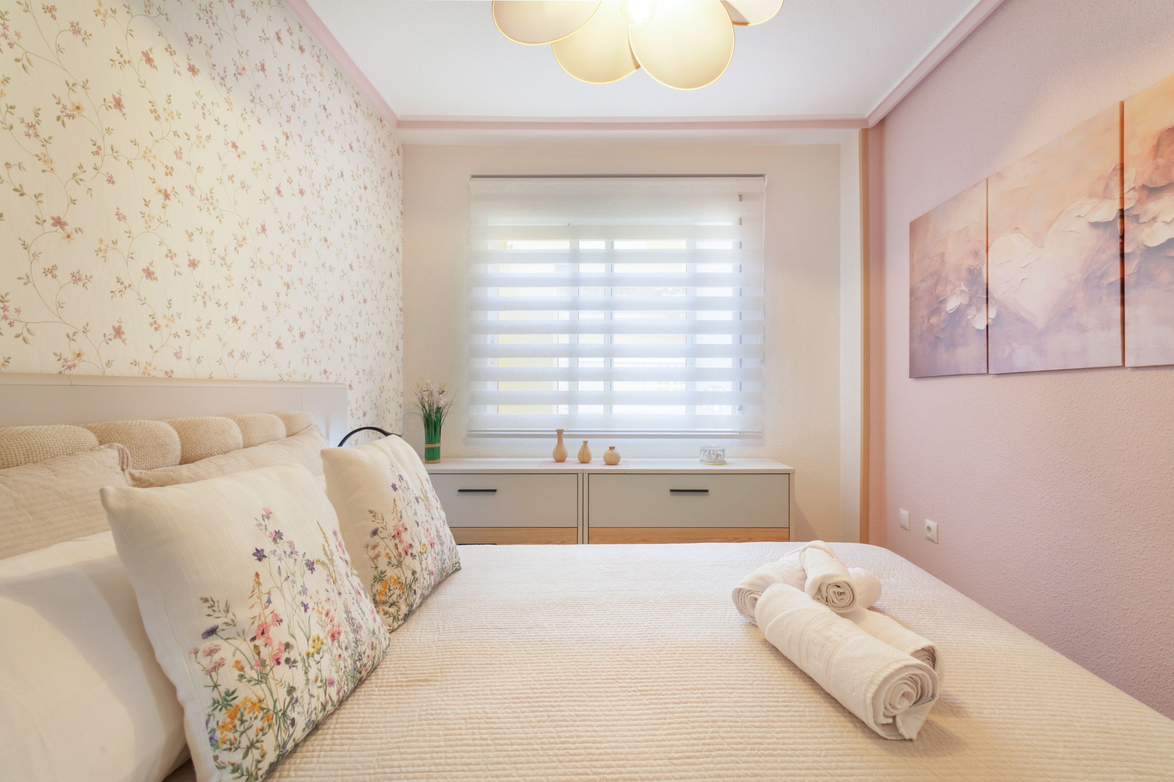 Un dormitorio con cama, cómoda, ventana con persianas y pared con estampado floral.