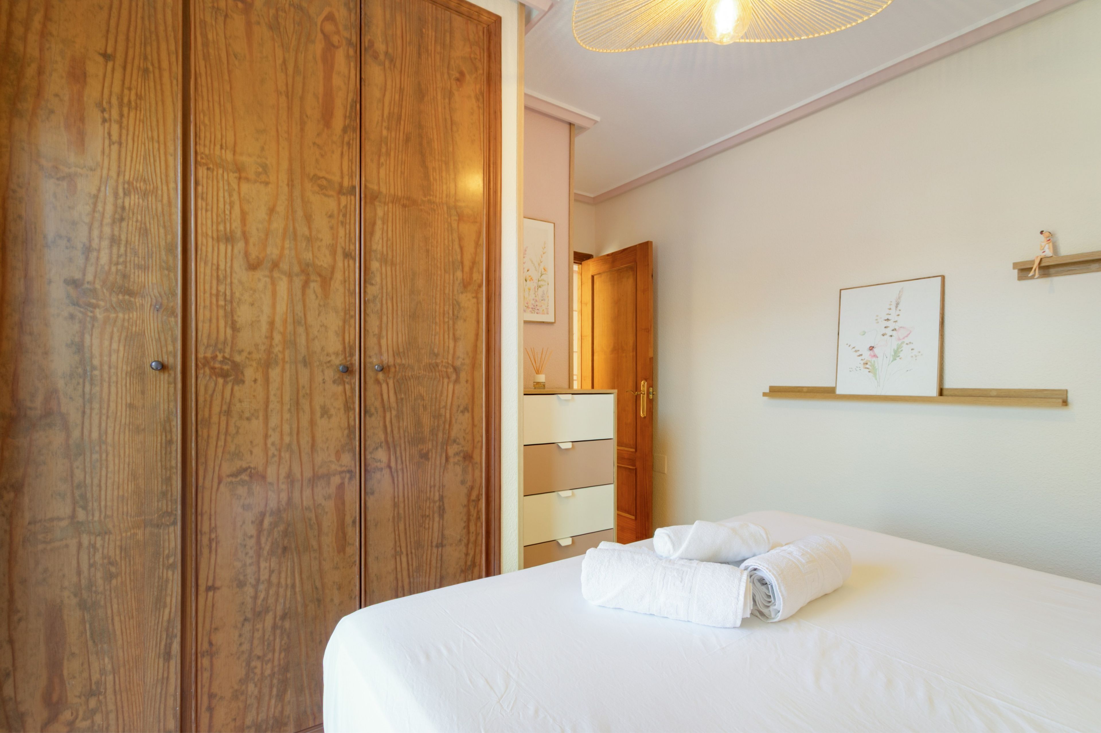 Un dormitorio con un amplio armario de madera, una cama con ropa de cama blanca y un estante de pared con un cuadro enmarcado.