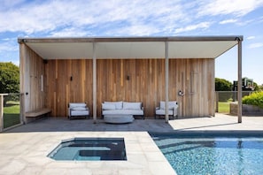 Un espace piscine extérieur moderne, doté d’une terrasse en bois, d’un coin salon couvert avec des canapés et d’un jacuzzi.