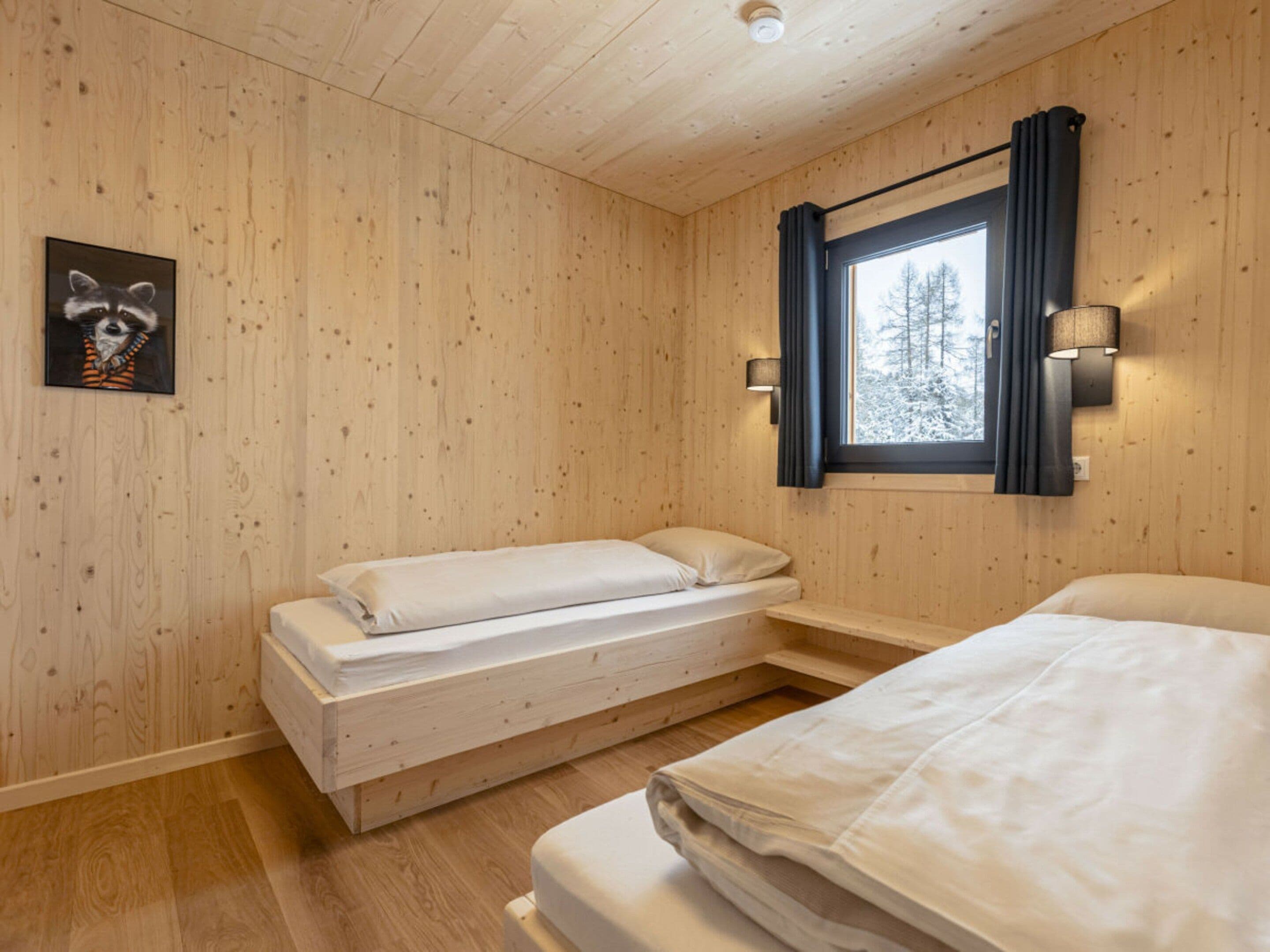 Una habitación con interior de madera, una cama, un banco, una ventana con cortinas y una lámpara fijada a la pared.