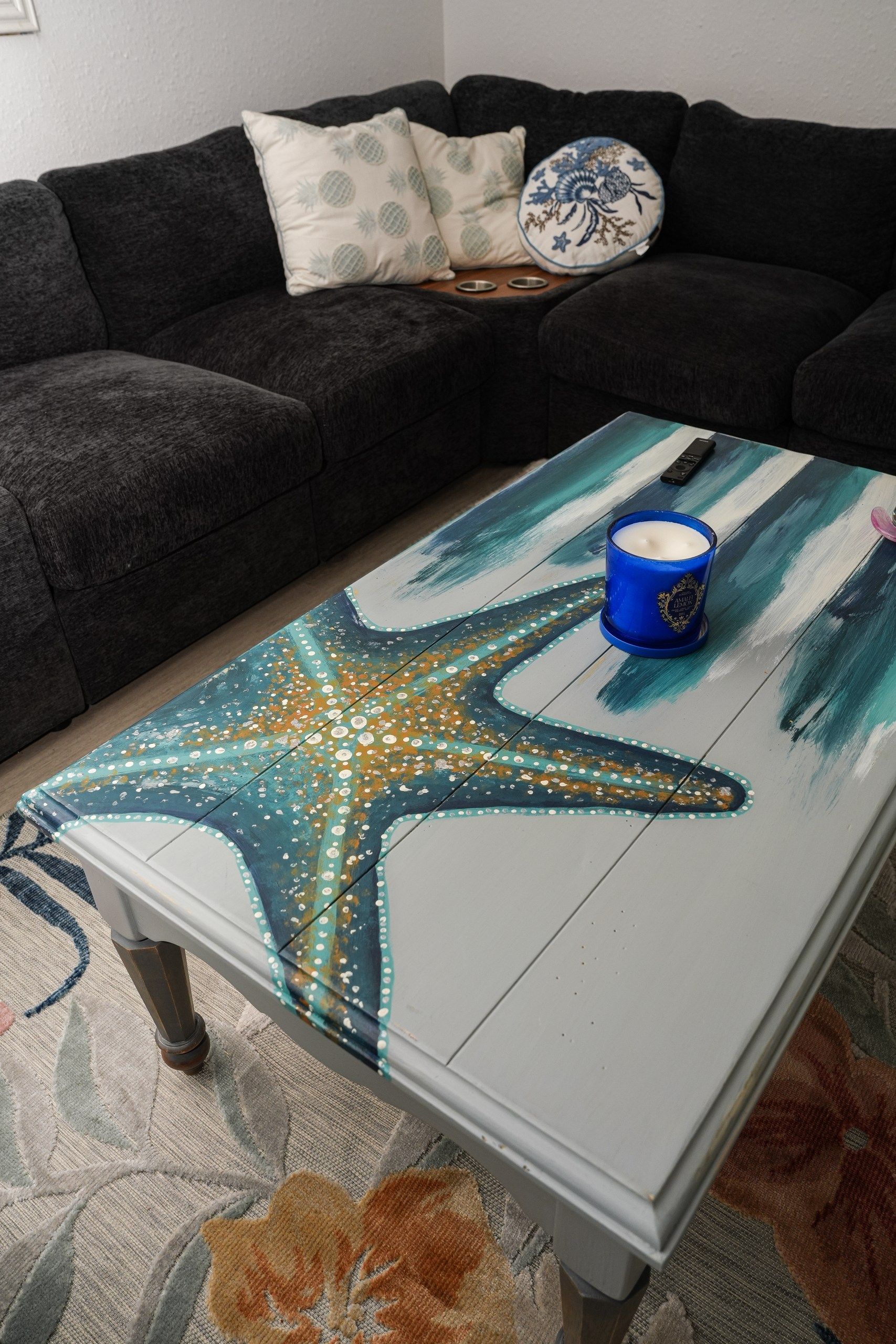 Una habitación con un sofá gris oscuro, una mesita de centro con un diseño de estrella de mar pintada y un candelabro azul.