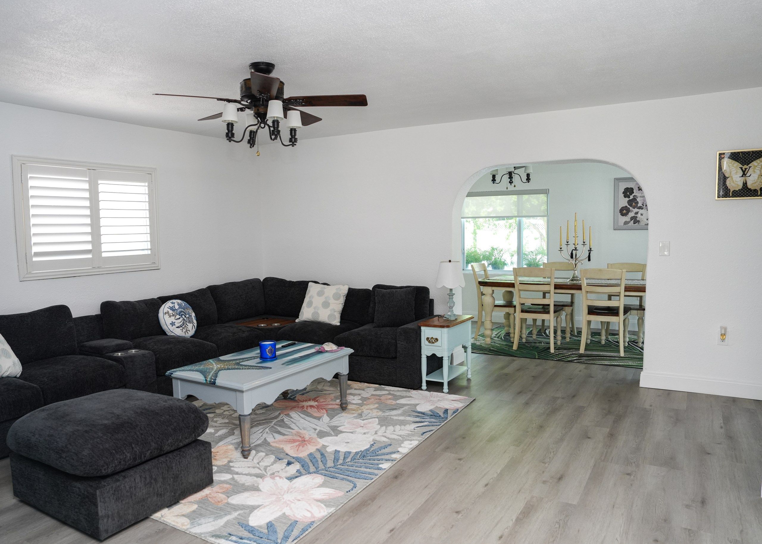 A living room with a dark gray sectional sofa, a coffee table with a floral rug, a ceiling fan, and a dining area with a wooden table and chairs.