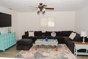 A living room with a sectional sofa, a coffee table, a teal chest of drawers, a television, and a ceiling fan.